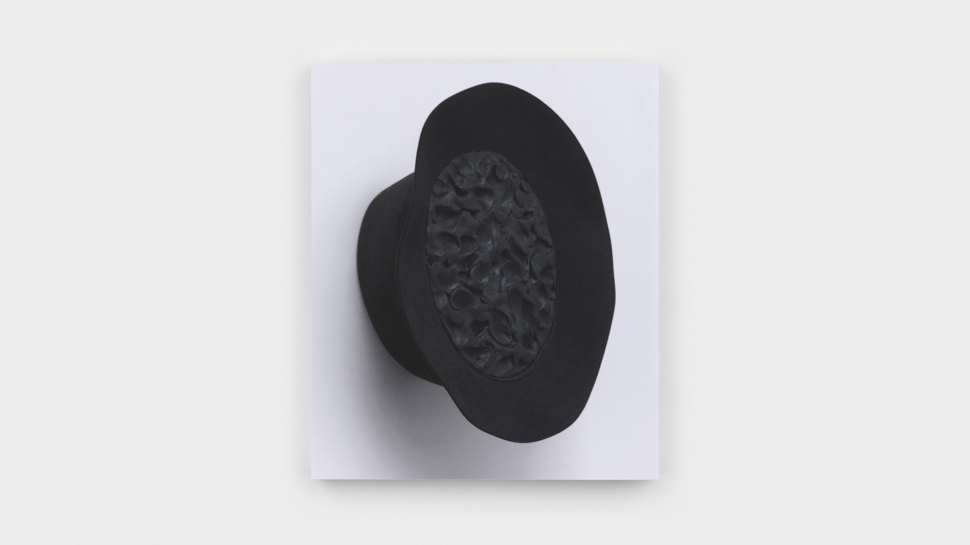 The book's back cover, featuring a black felt fedora affixed to the wall. Its cavity is filled with dark clay pocked with a finger's pinches.