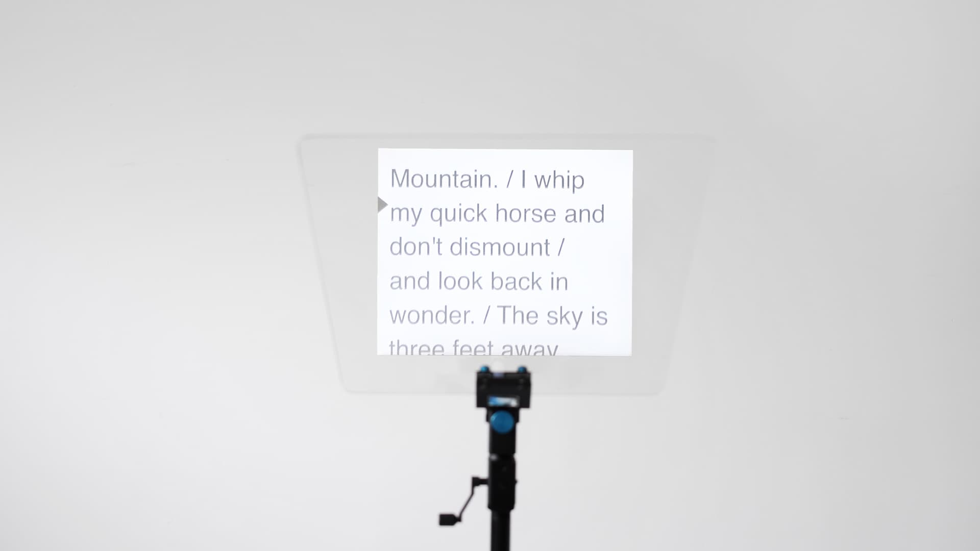 A close-up, front-on photograph of the teleprompter's glass panel. The image projected on to the screen is now visible, revealing large black sans-serif text that reads: 'Mountain./I whip my quick horse and do not dismount / and look back in wonder. / The sky is three feet away.'