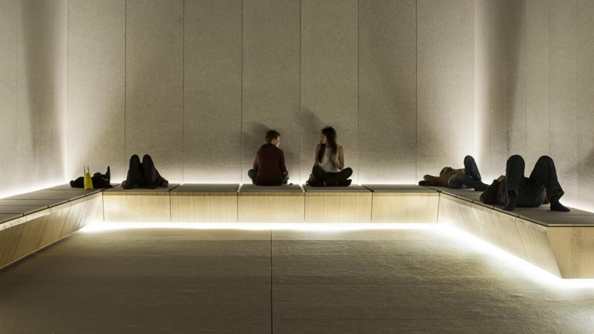 Selfridge's austere Silence Room, with padded beige felt walls and light wood benches running its circumference. Two shoppers lay face-up, while two children sit cross-legged, one facing the wall.