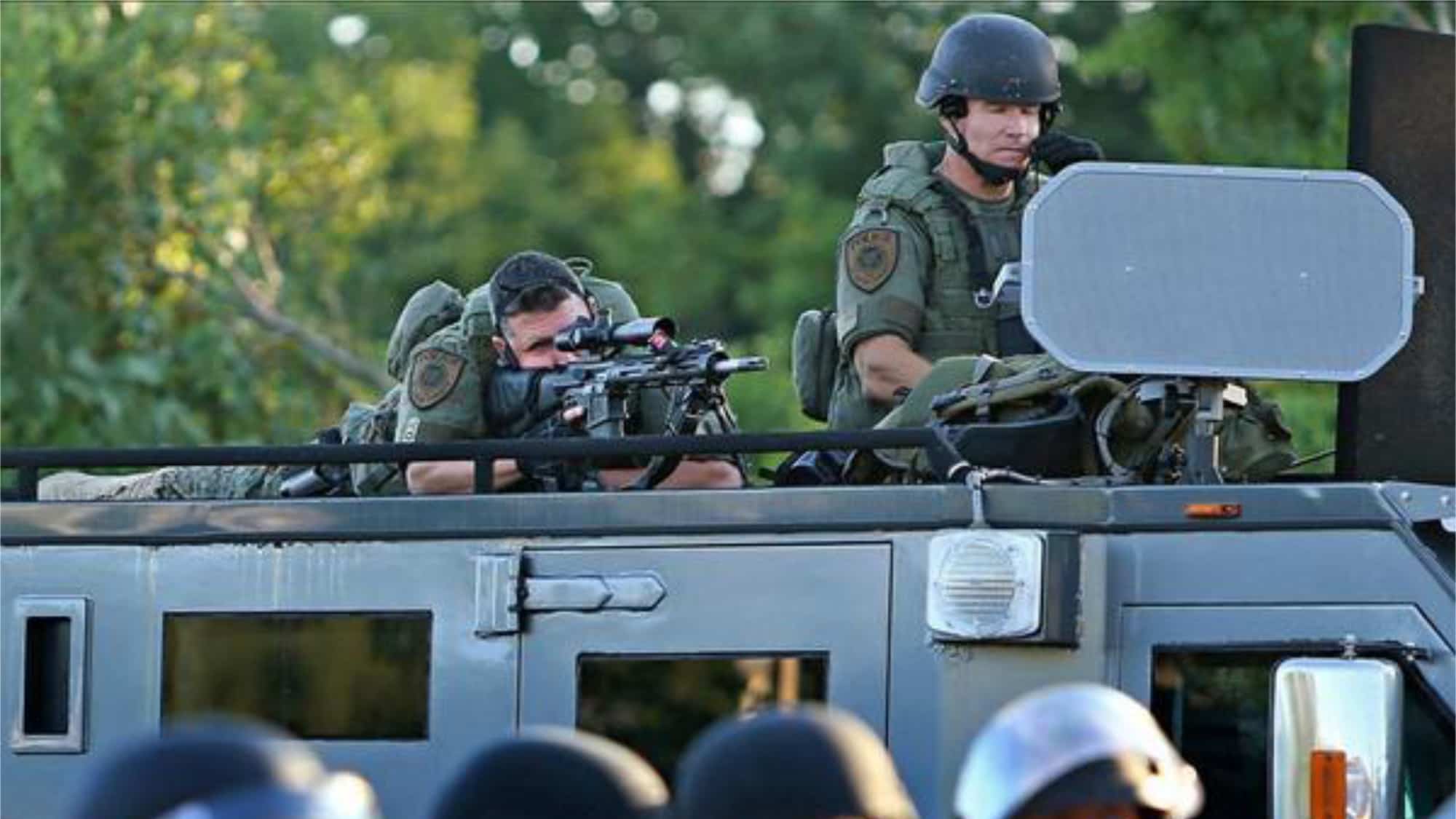 Two white male police officers dressed in tactical gear atop an armored vehicle. The officer at right speaks into a microphone attached to his black helmet, which broadcasts through the rectangular grey speaker in front of him. The officer to his left lies on his stomach and looks through the scrope of an automatic rifle, with his finger on the trigger. 